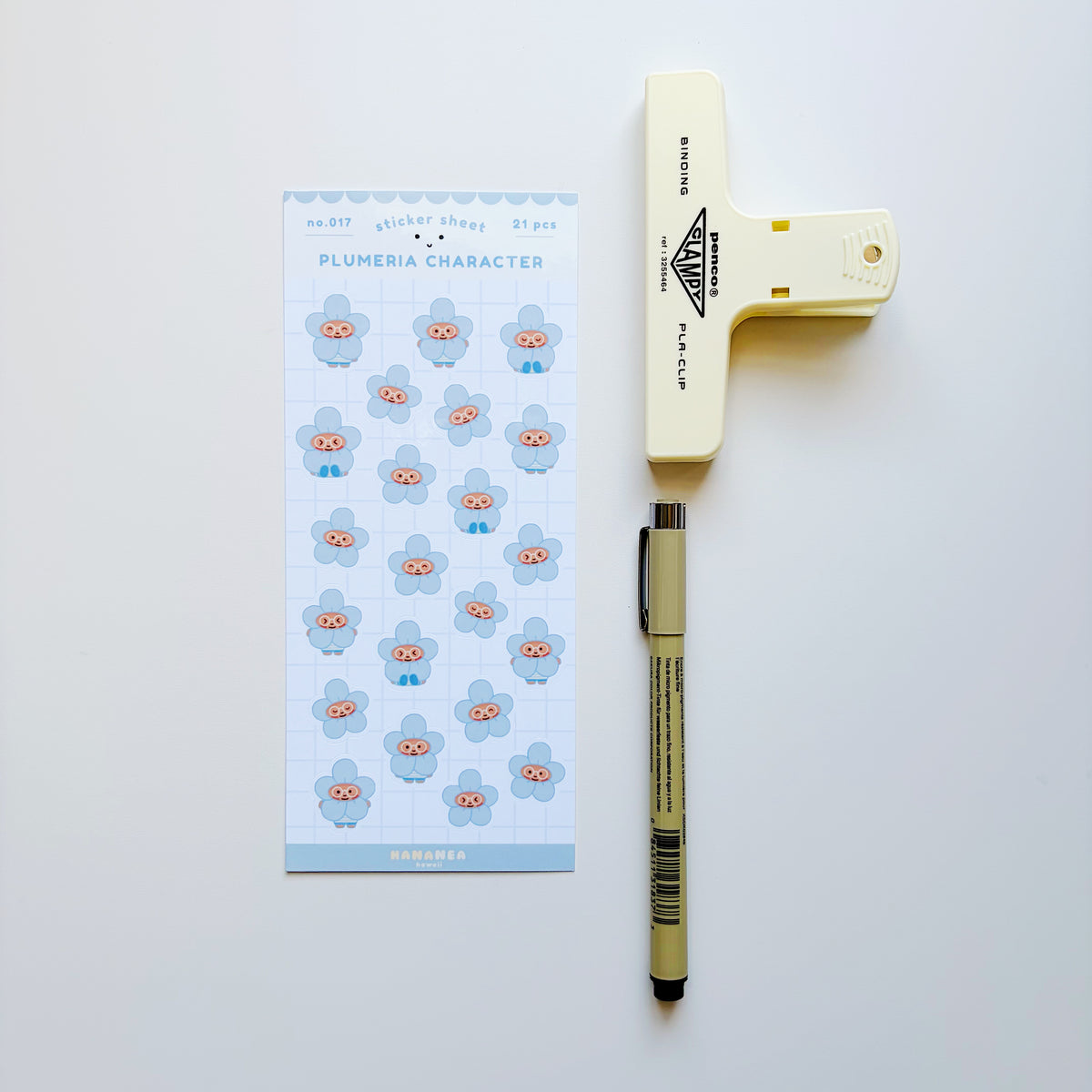 A sheet of planner stickers featuring cute plumeria flower characters, displayed next to a cream-colored paper clip and a black pen on a clean white background.