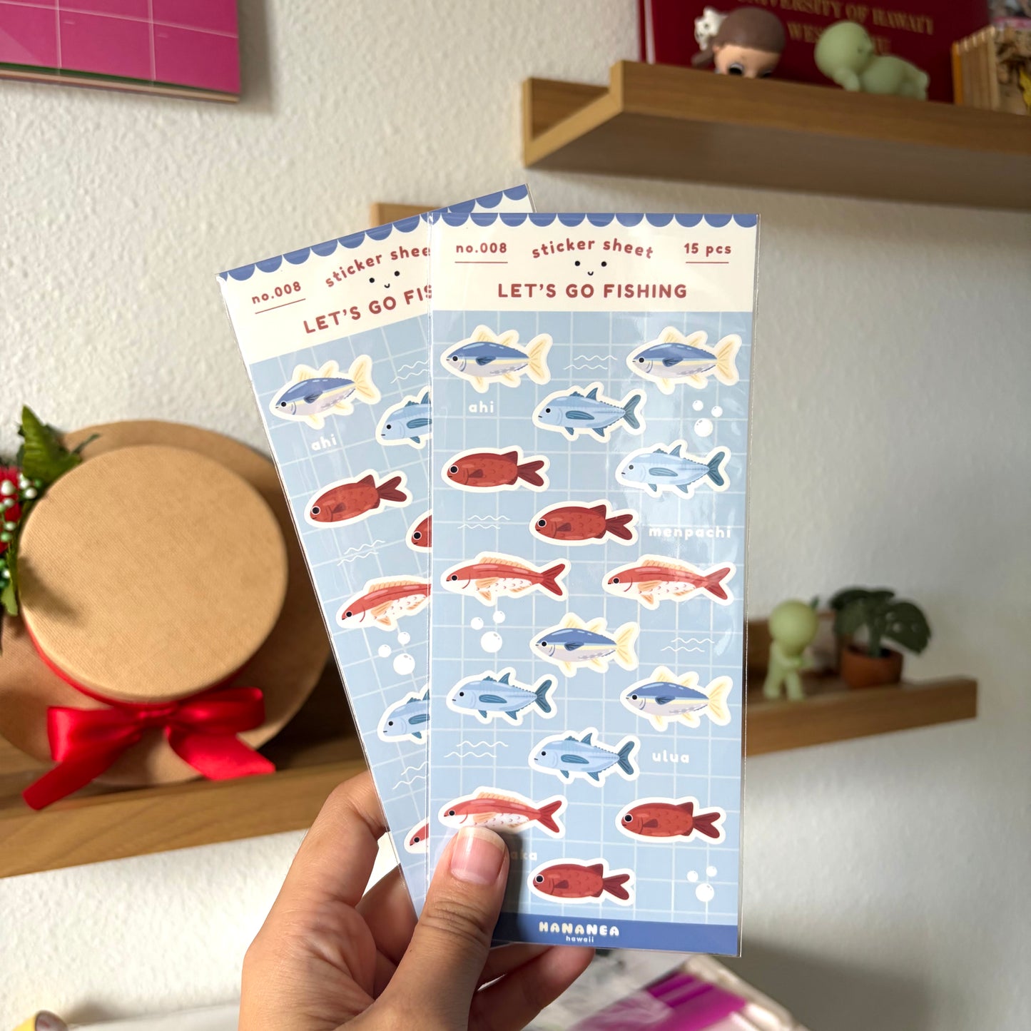 Two sticker sheets with fish designs held by a hand against a home decor background.