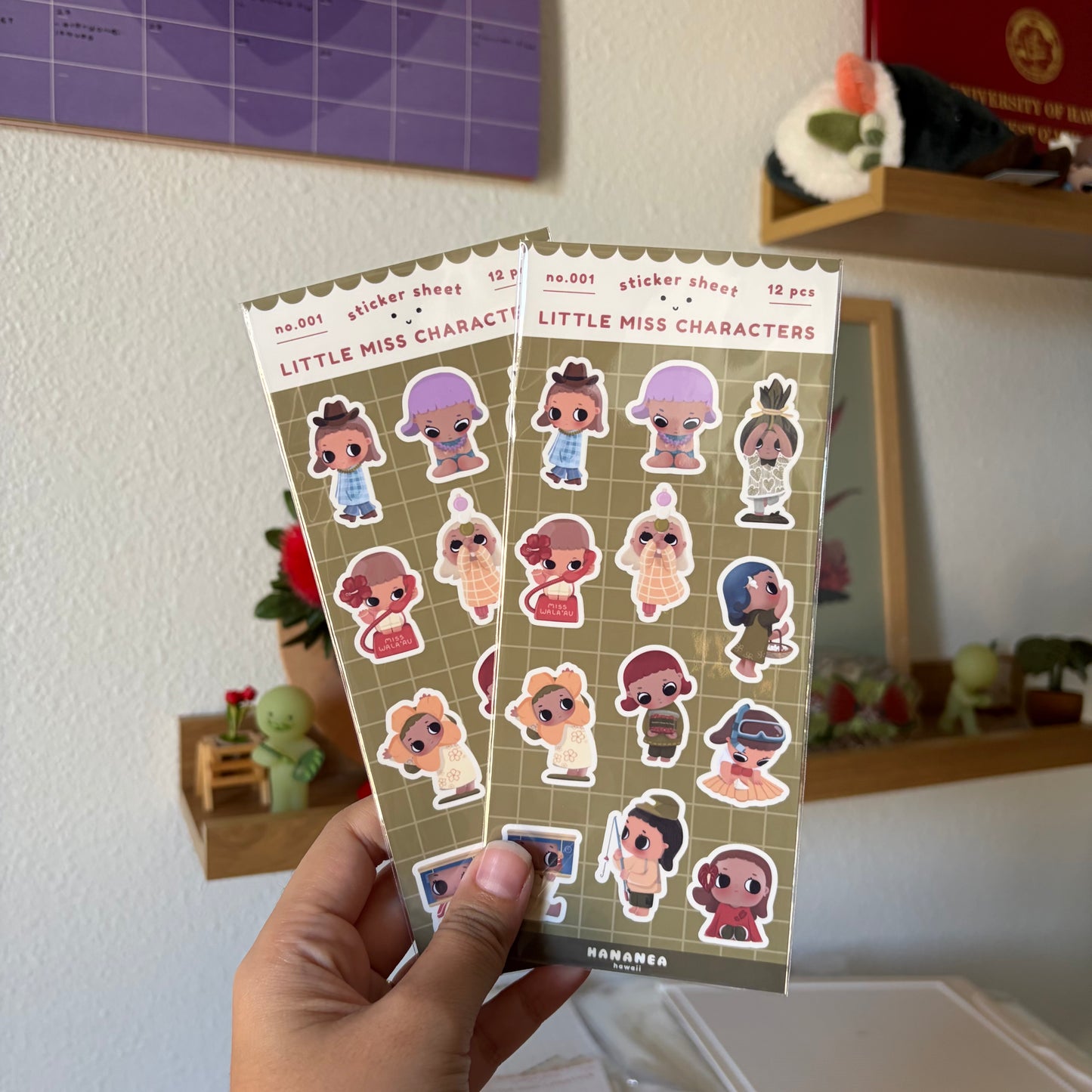 A hand holds two sticker sheets against a blurred indoor room background.