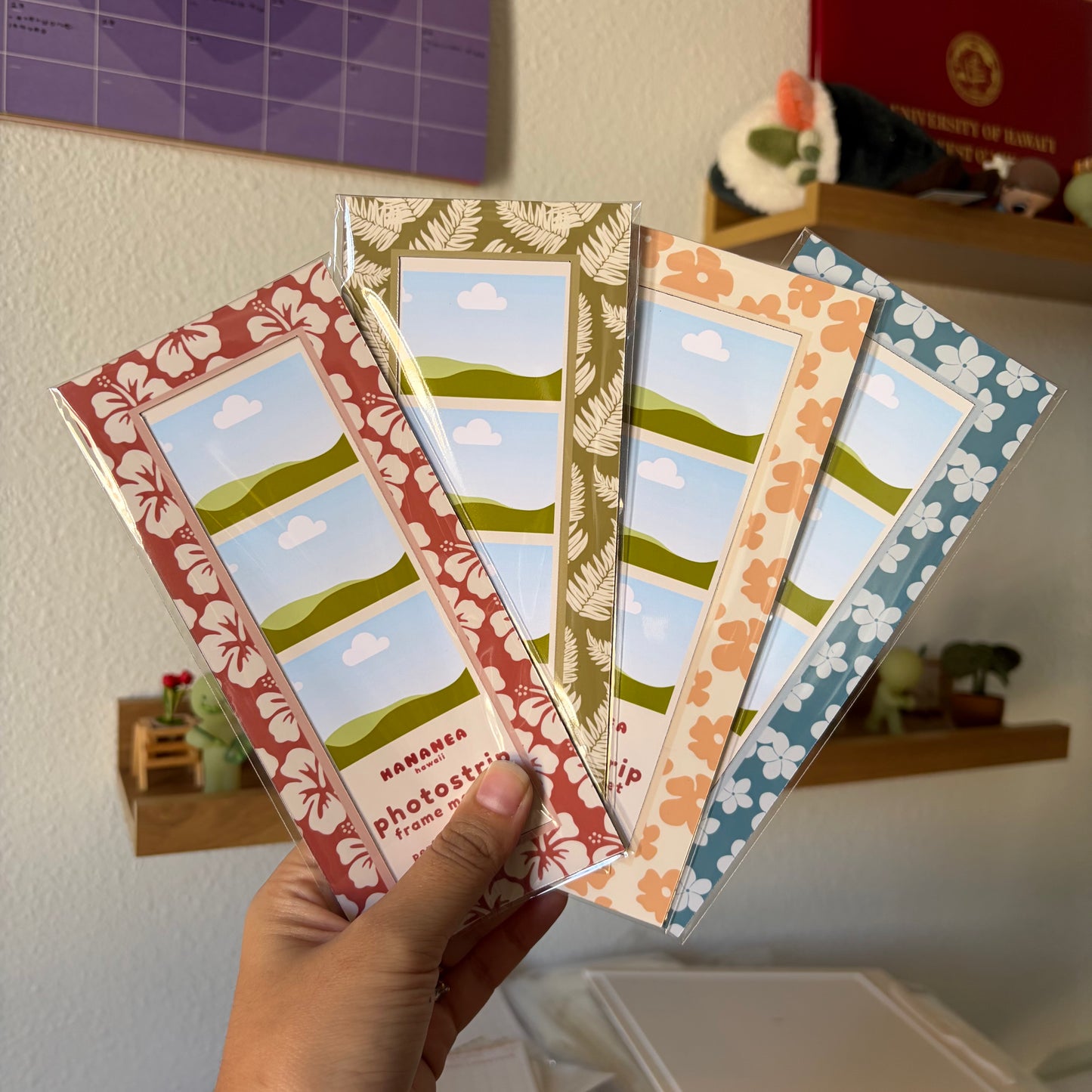 A hand holds four rectangular photo strip frames with tropical floral borders in red, olive green, peach, and blue, each featuring a landscape illustration of green hills under a blue sky with white clouds.