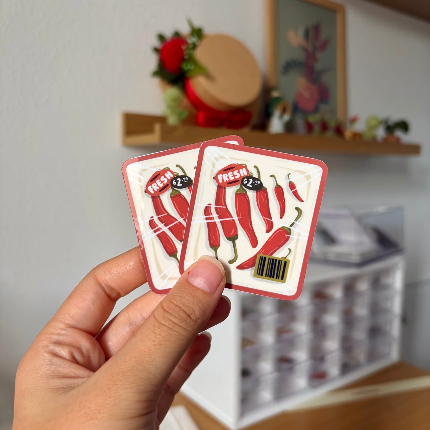 Hand holding two stickers with red chili pepper design in a room setting.