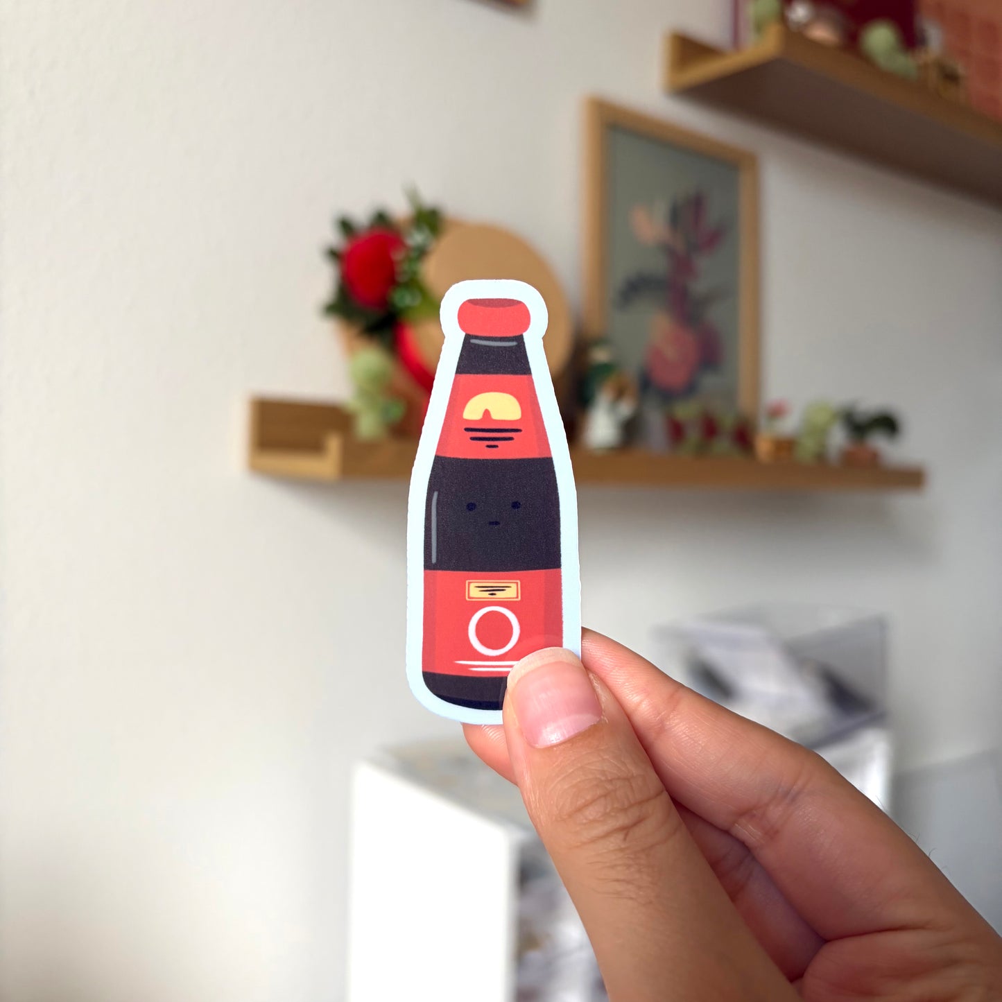 Hand holding a red and black bottle-shaped sticker with a blurred indoor background