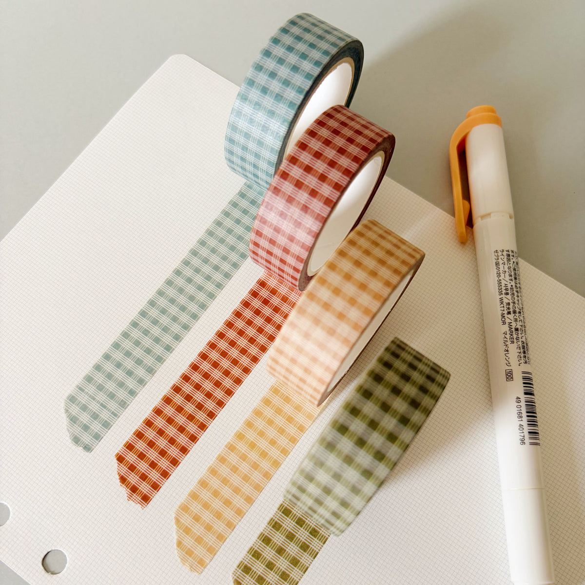 Four rolls of palaka washi tape in pastel shades of blue, red, yellow, and green, arranged on a white notebook page next to a white highlighter pen with an orange cap.