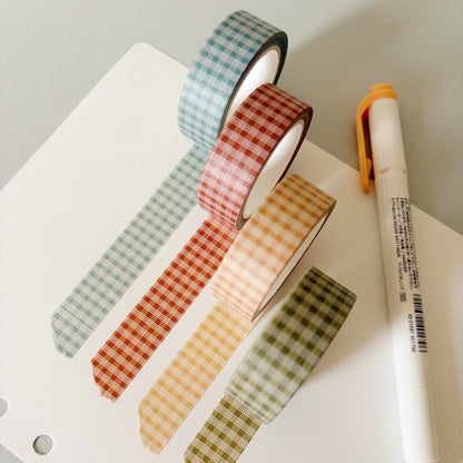 Four rolls of palaka washi tape in pastel shades of blue, red, yellow, and green, arranged on a white notebook page next to a white highlighter pen with an orange cap.