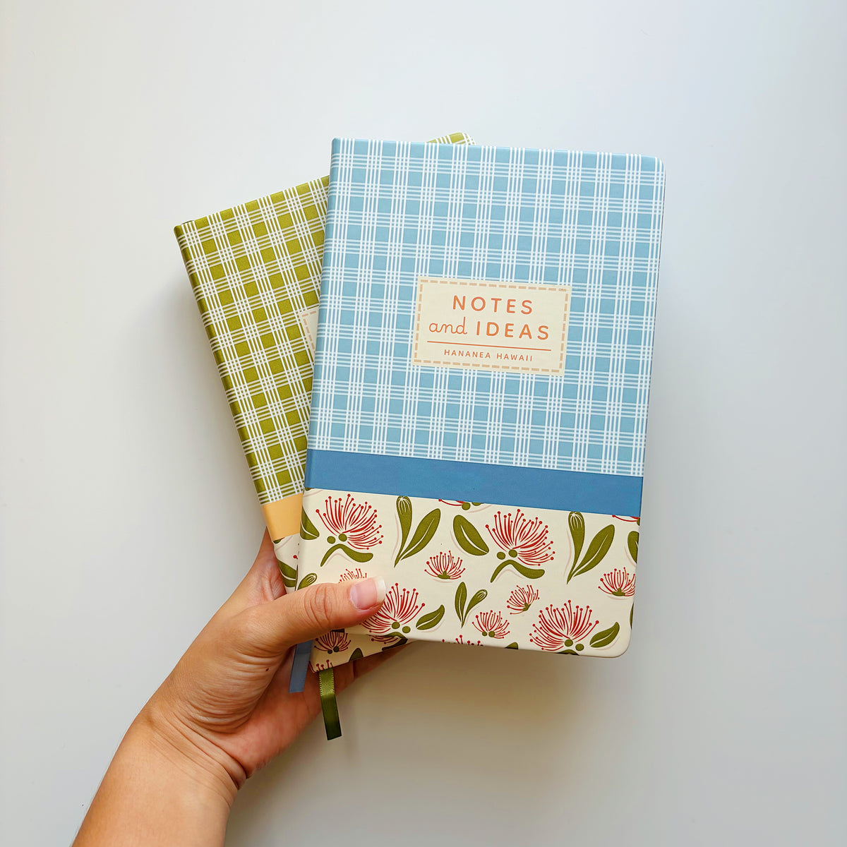 A hand holds two floral and palaka patterned notebooks against a plain light background.