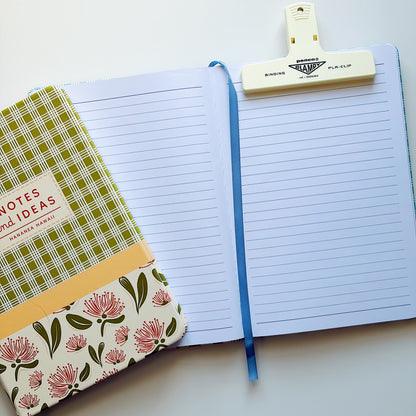An open lined notebook with a blue ribbon bookmark, held by a cream-colored pen clip. A patterned notebook cover with pink flowers and green leaves rests on the left side.