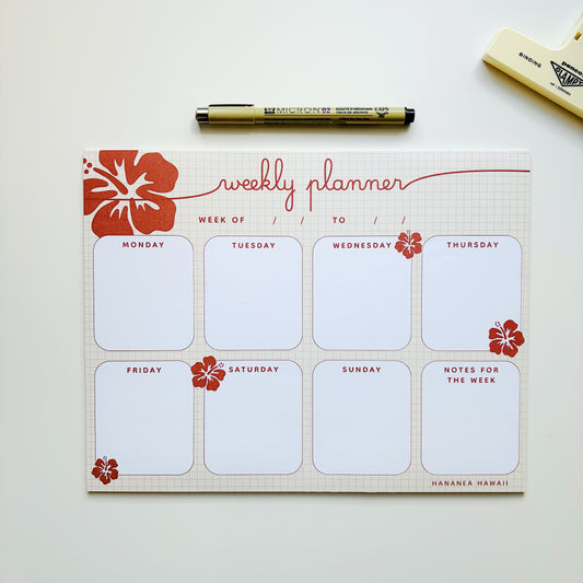 Red weekly planner notepad with red flowers and a tan pen.
