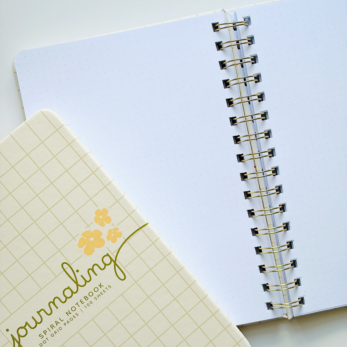 A close-up of a spiral-bound notebook with a white page and gold-toned metal rings, alongside a closed matching notebook cover in pale yellow with a grid pattern and the word 'journaling' in gold script.