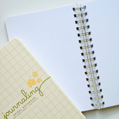 A close-up of a spiral-bound notebook with a white page and gold-toned metal rings, alongside a closed matching notebook cover in pale yellow with a grid pattern and the word 'journaling' in gold script.