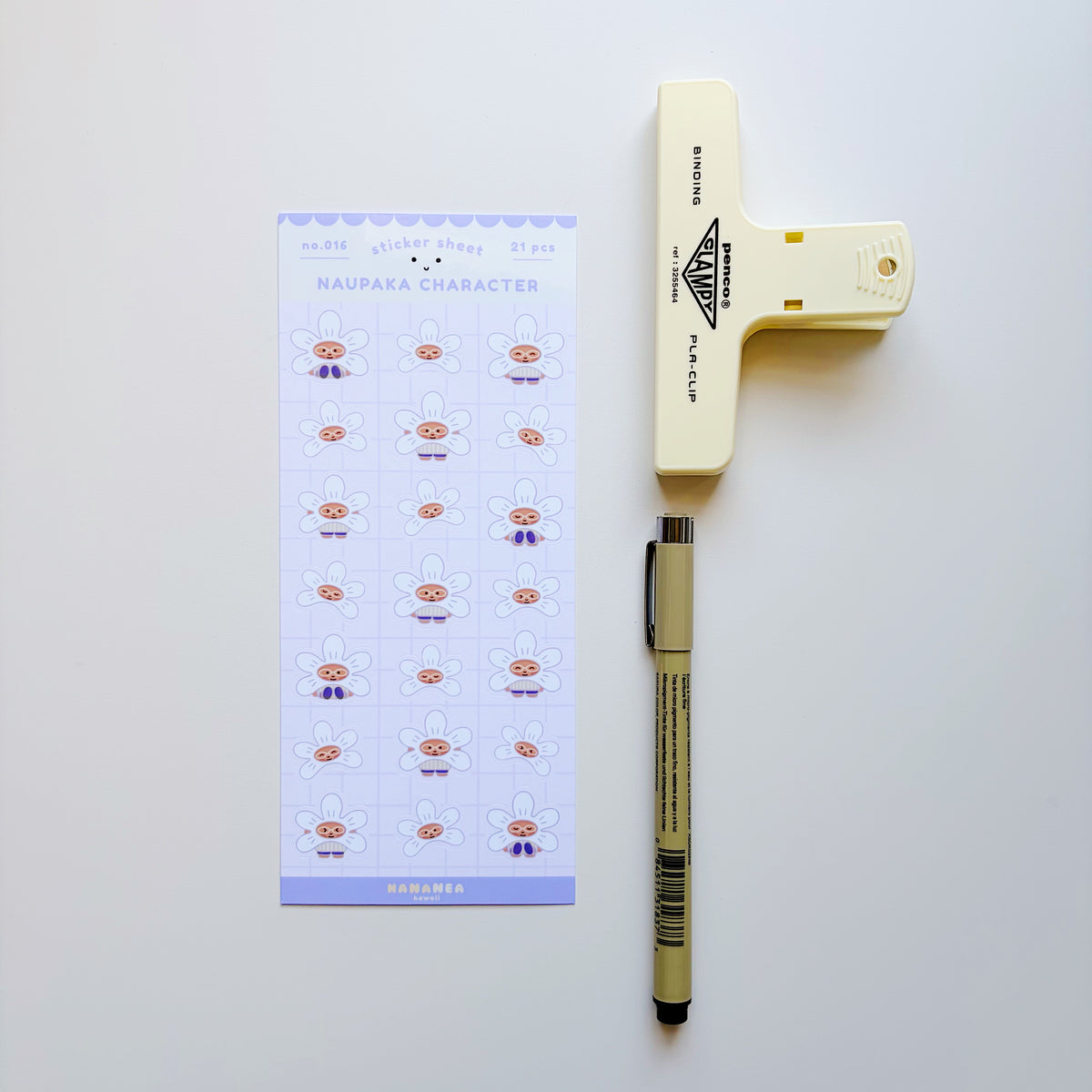 A flat-lay of stationery items on a white background: a printed sheet labeled 'NAUPAKA CHARACTER' with small illustrated character stickers, a cream-colored paper clip, and a beige pen.