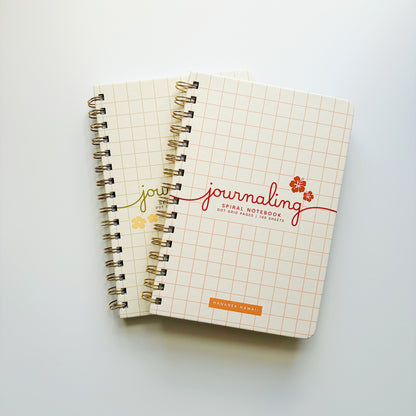 Two spiral-bound notebooks with a grid pattern cover, stacked on a light gray background. The top notebook features the word 'journaling' in red script with small red flowers.