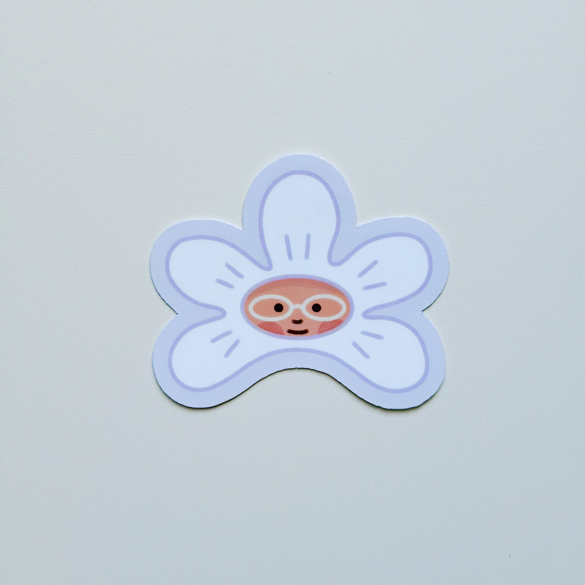 A die-cut sticker of a smiling cartoon flower with a face peeking out from the center, shown on a plain light gray background.