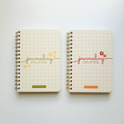 Two spiral-bound notebooks with cream grid covers are displayed side by side on a light gray background. The left notebook features a green 'journaling' script with yellow floral accents, and the right notebook features a red 'journaling' script with orange floral accents.