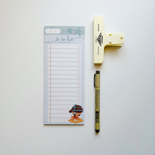 A flat-lay of a beach-themed to-do list notepad, a cream-colored paper clip, and a tan and black pen on a clean white background.