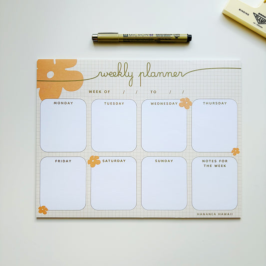 A weekly planner notepad with a grid background, orange floral accents, and sections for Monday through Sunday plus notes for the week, styled on a light surface with a black pen and a small yellow item nearby.