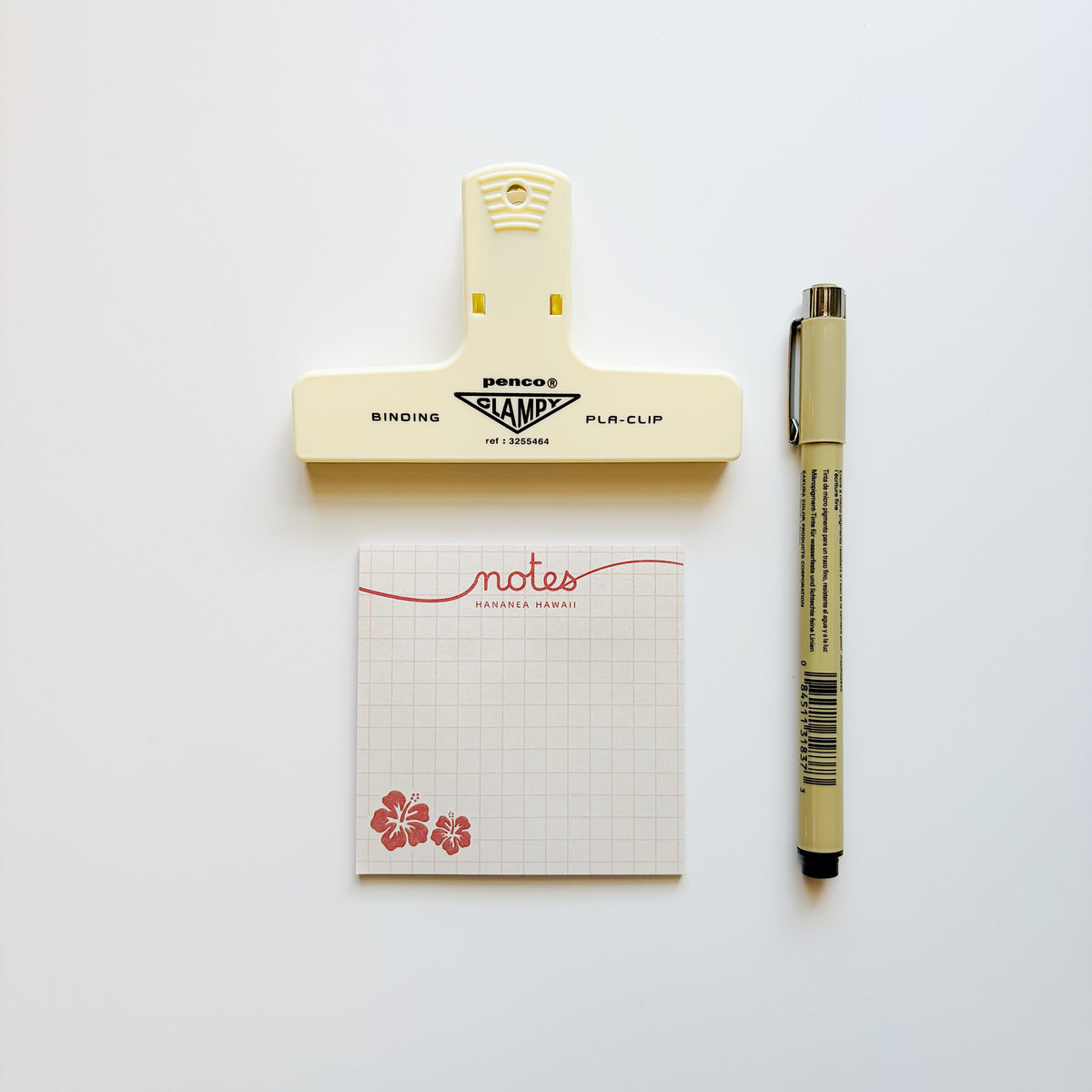 A flat-lay of stationery items on a white background: a cream-colored binding stamp clip, a small grid notepad with red script and floral decoration, and a beige pen.