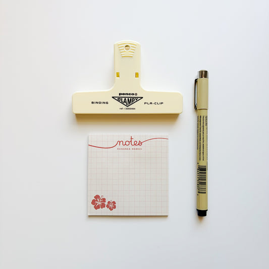 A flat-lay of stationery items on a white background: a cream-colored binding stamp clip, a small grid notepad with red script and floral decoration, and a beige pen.