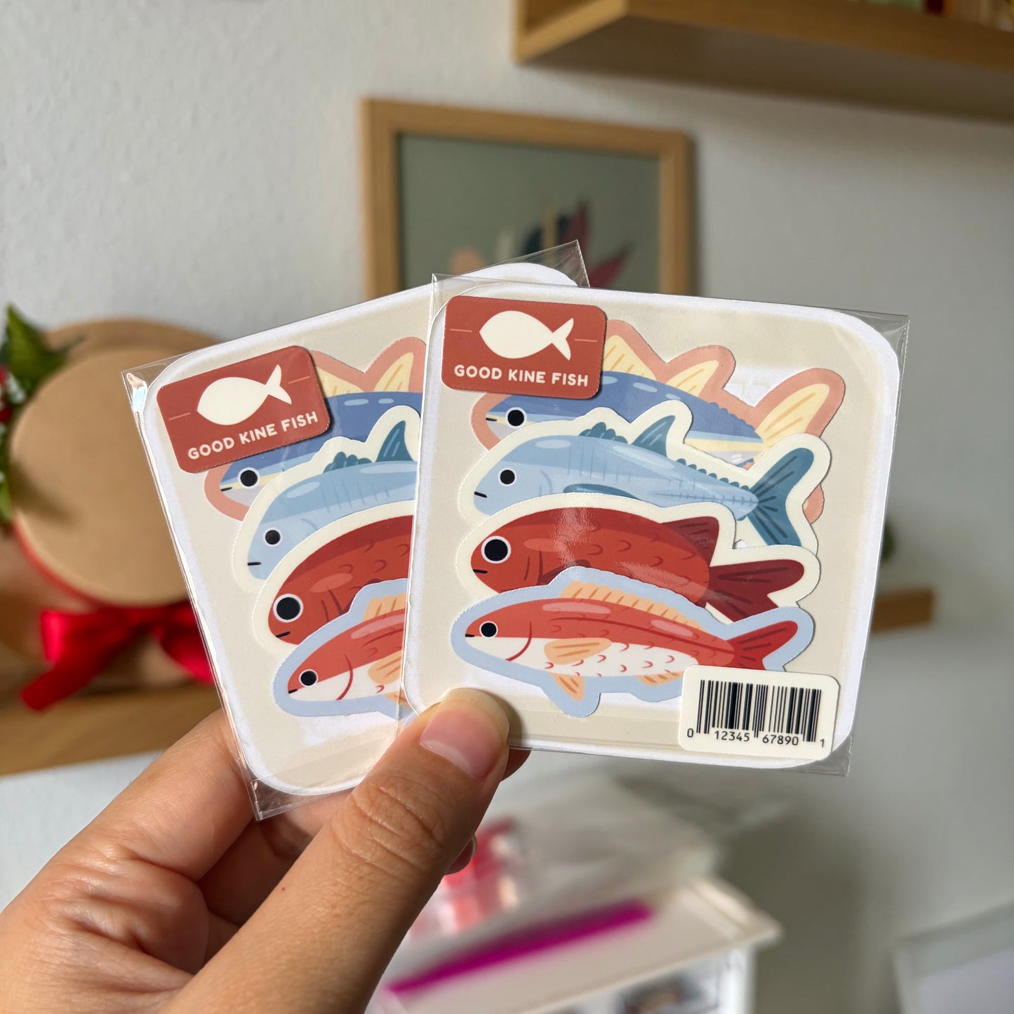 Hand holding two packages of colorful fish stickers with a blurred indoor background.