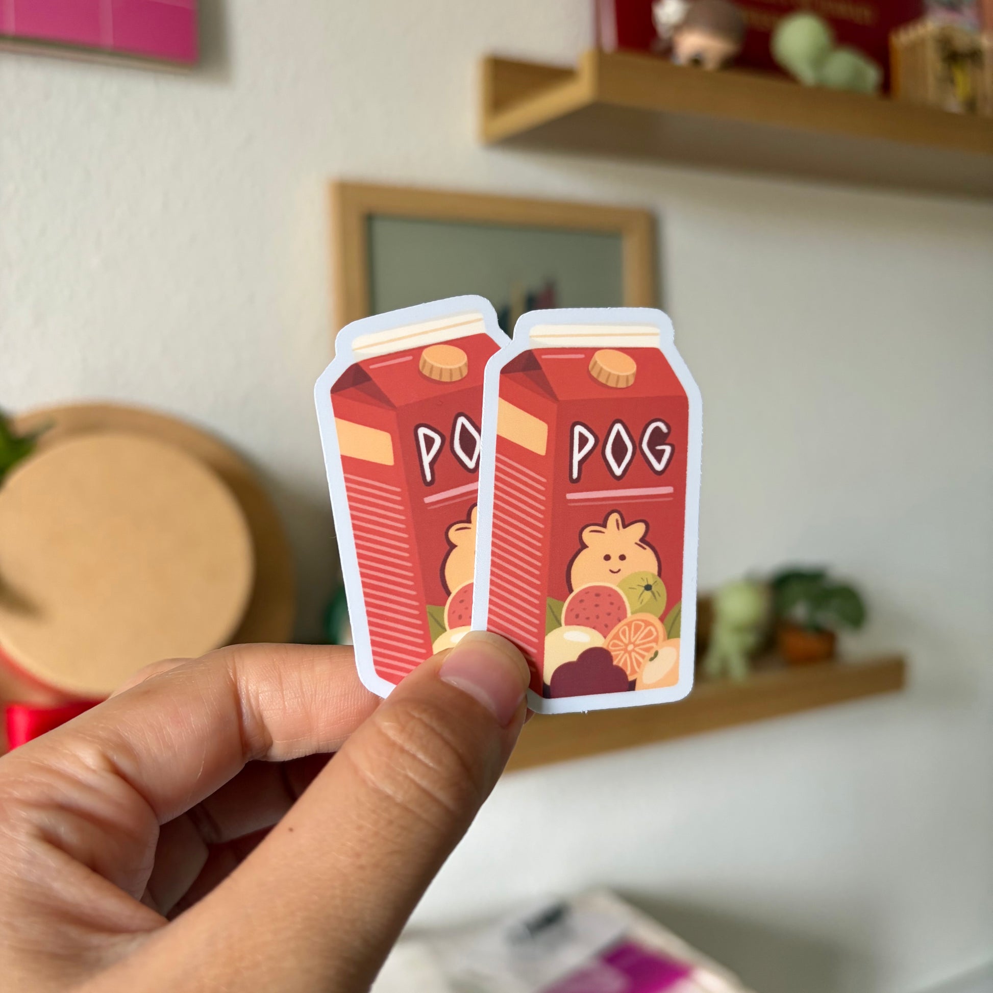 Two small red POG stickers held by a hand in front of a blurred indoor background.