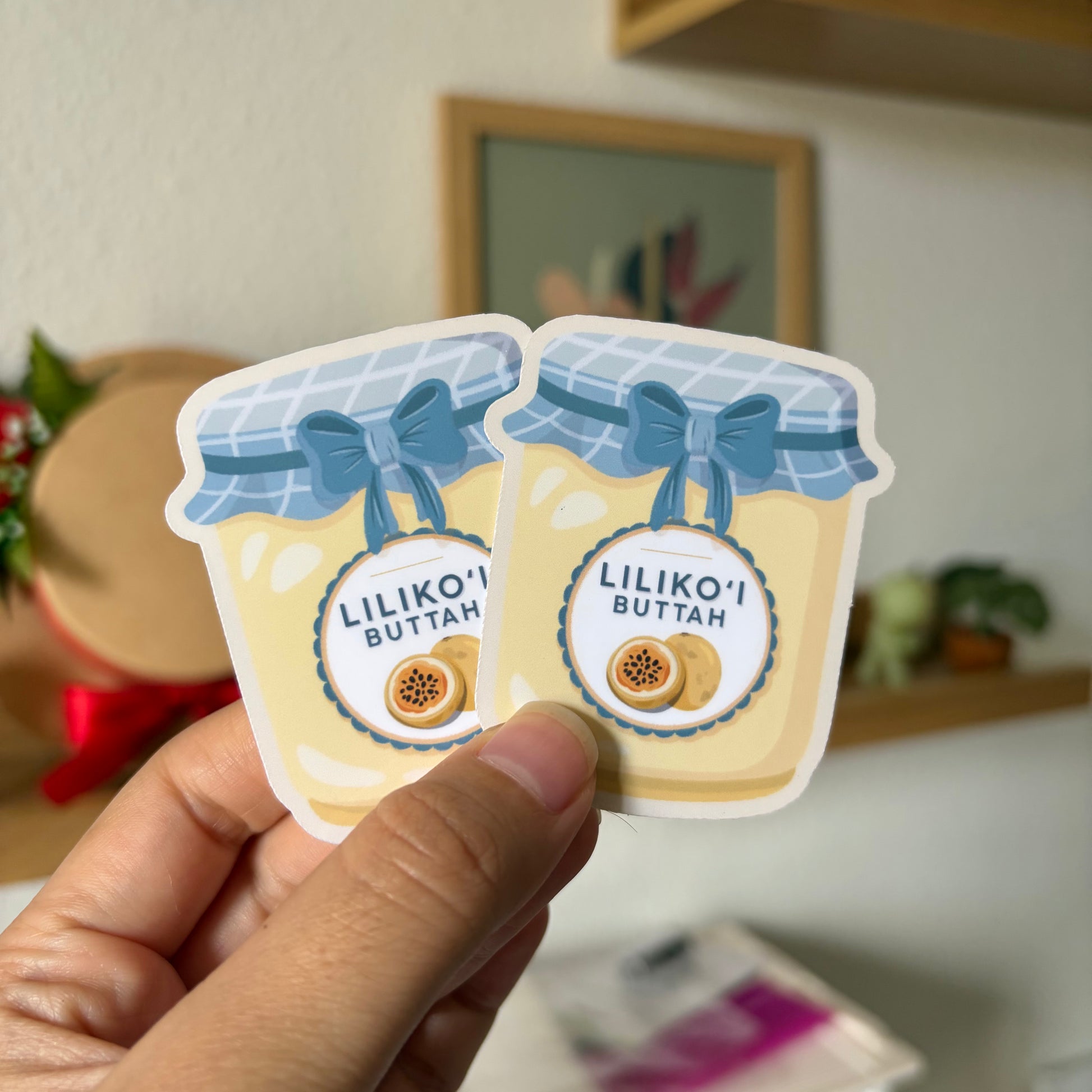 Two stickers of 'Lilikoi Buttah' in a hand with a blurred indoor background