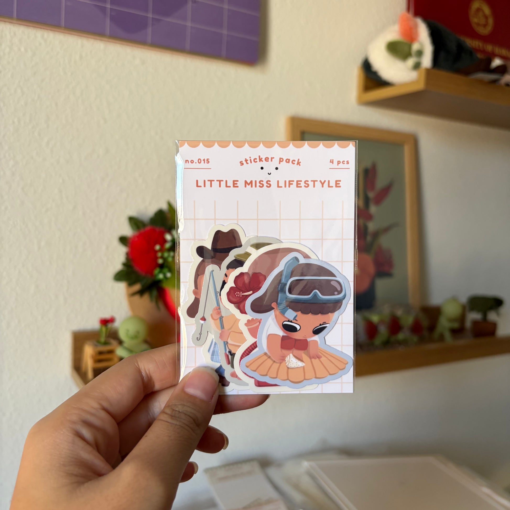 A hand holds a sticker pack labeled 'LITTLE MISS LIFESTYLE' in a home setting with shelves and framed art in the background.