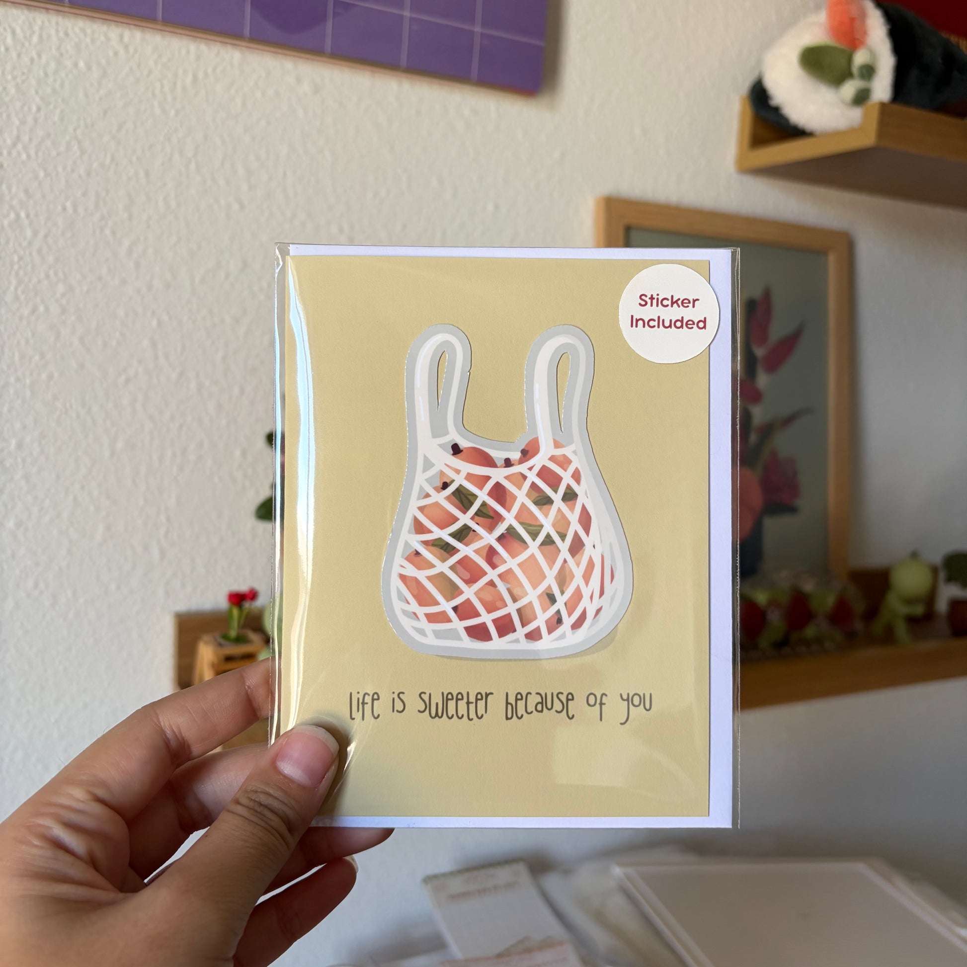 A hand holds a square greeting card featuring a reusable mesh bag illustration with the text 'life is sweeter because of you' and a small 'Sticker Included' badge. The background shows a softly blurred indoor room with shelves and decor.