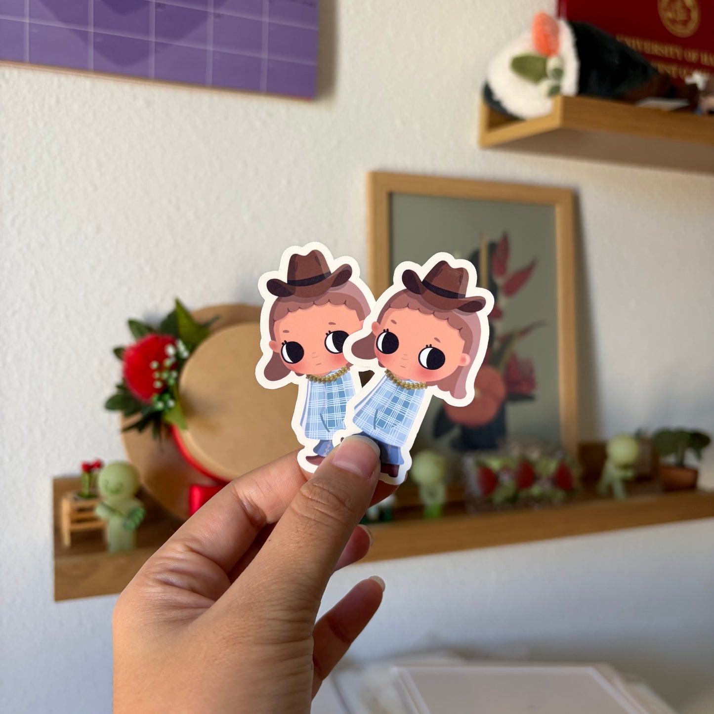 A hand holds a die-cut sticker featuring two cartoons.