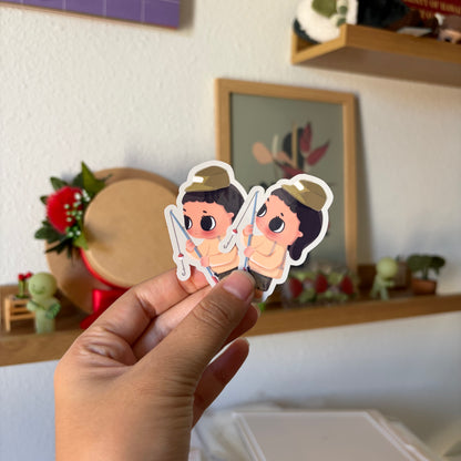 A hand holds two small die-cut stickers featuring cartoon characters.