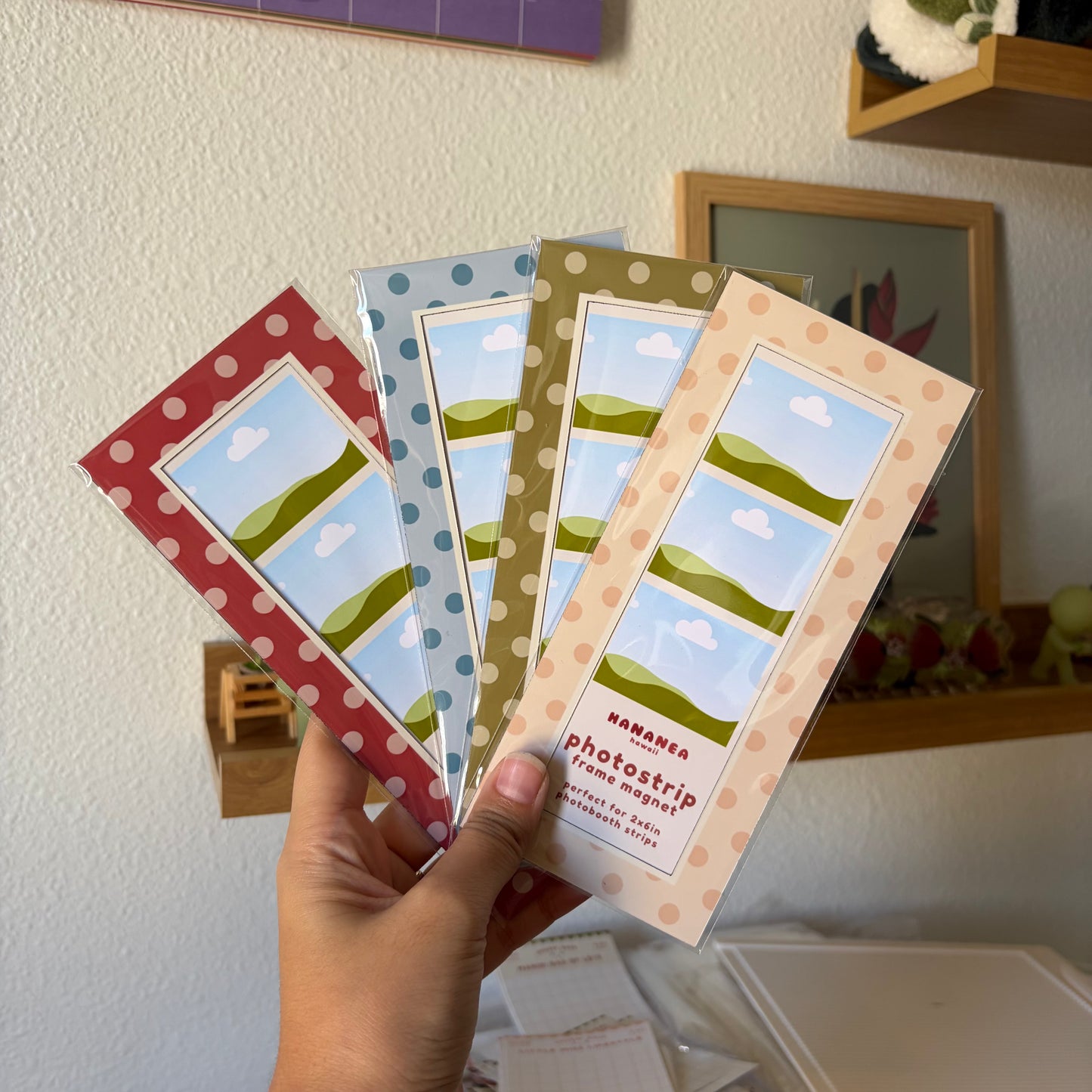 A hand holds four photo strips in a fan arrangement, each with a different pastel color border (red, blue, olive green, and peach) with white polka dots and a central landscape illustration of green hills under a blue sky with white clouds.