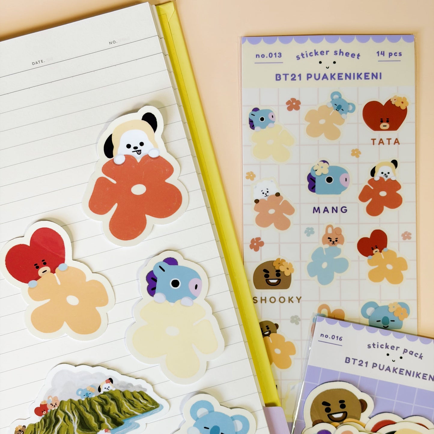 A styled flat-lay of BT21 Puaenikeni sticker sheets and individual die-cut stickers arranged on a lined notebook page next to a yellow pencil, on a peach background.