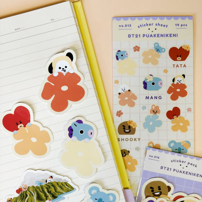 A styled flat-lay of BT21 Puaenikeni sticker sheets and individual die-cut stickers arranged on a lined notebook page next to a yellow pencil, on a peach background.
