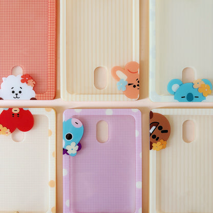 Six pastel phone cases arranged in a 2x3 grid on a pale pink background. Each case has a different color and pattern, including coral grid, cream stripes, pale yellow, lavender grid, cream with dots, and light blue, and each features a small cartoon character charm peeking out from the lower left corner.
