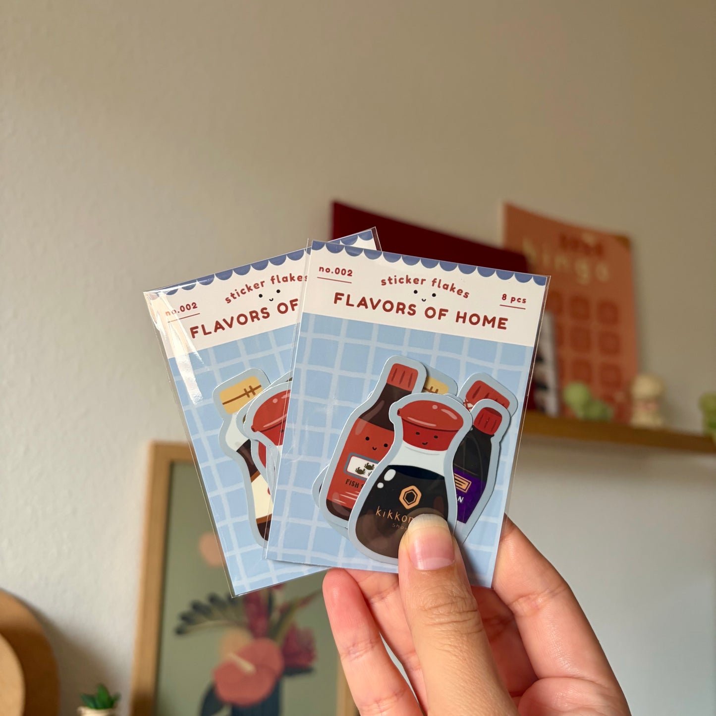 Two 'Flavors of Home' sticker flake packs held by a hand with a neutral background.