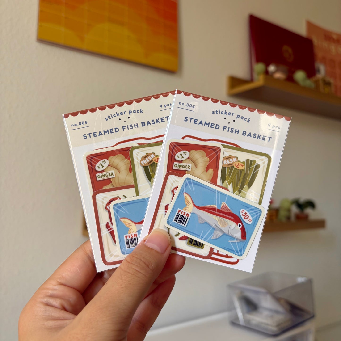 Two sticker packs labeled 'Steamed Fish Basket' held by a hand against a neutral background.