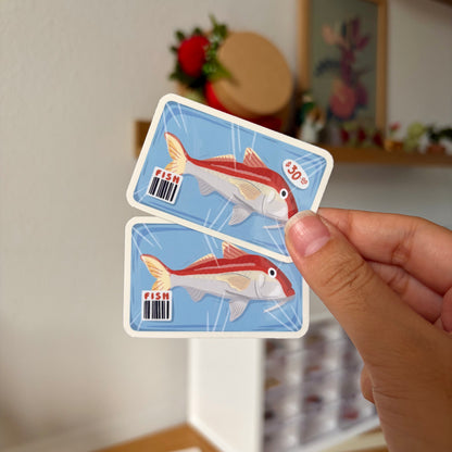 Hand holding two fish-themed stickers with a blurred background