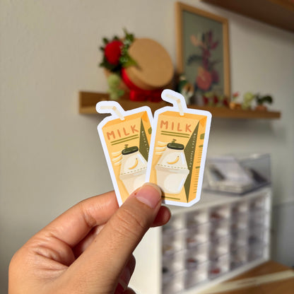 Two small stickers of milk cartons held by a hand with a blurred indoor background.