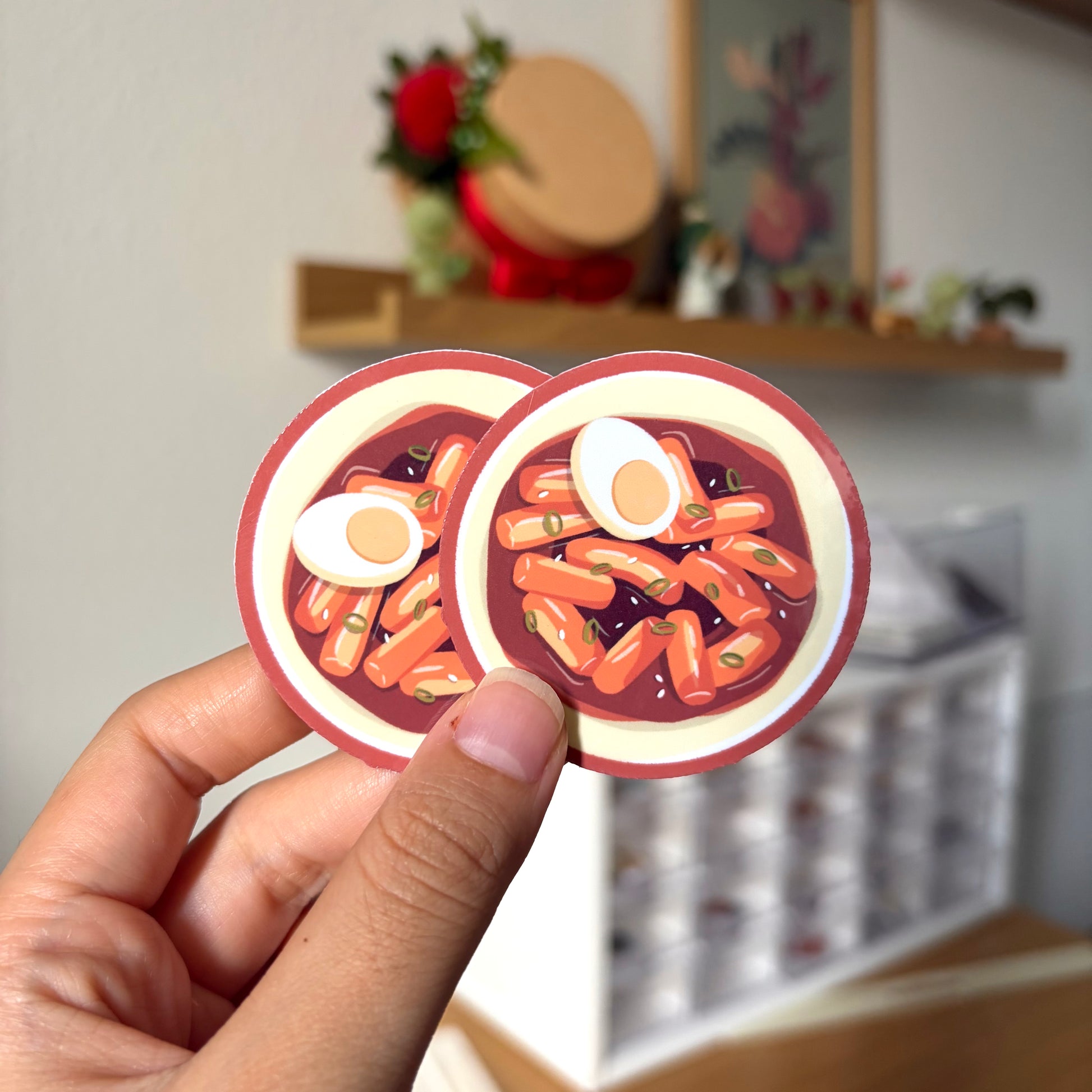 Two round stickers of tteokbokki with eggs held by a hand against a blurred indoor background.