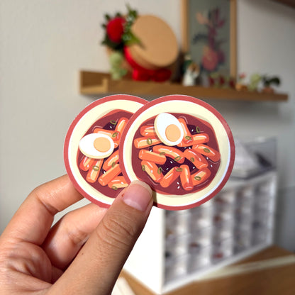 Two round stickers of tteokbokki with eggs held by a hand against a blurred indoor background.