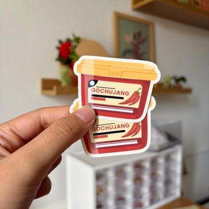 Sticker of a gochujang container held by a hand with a blurred indoor background