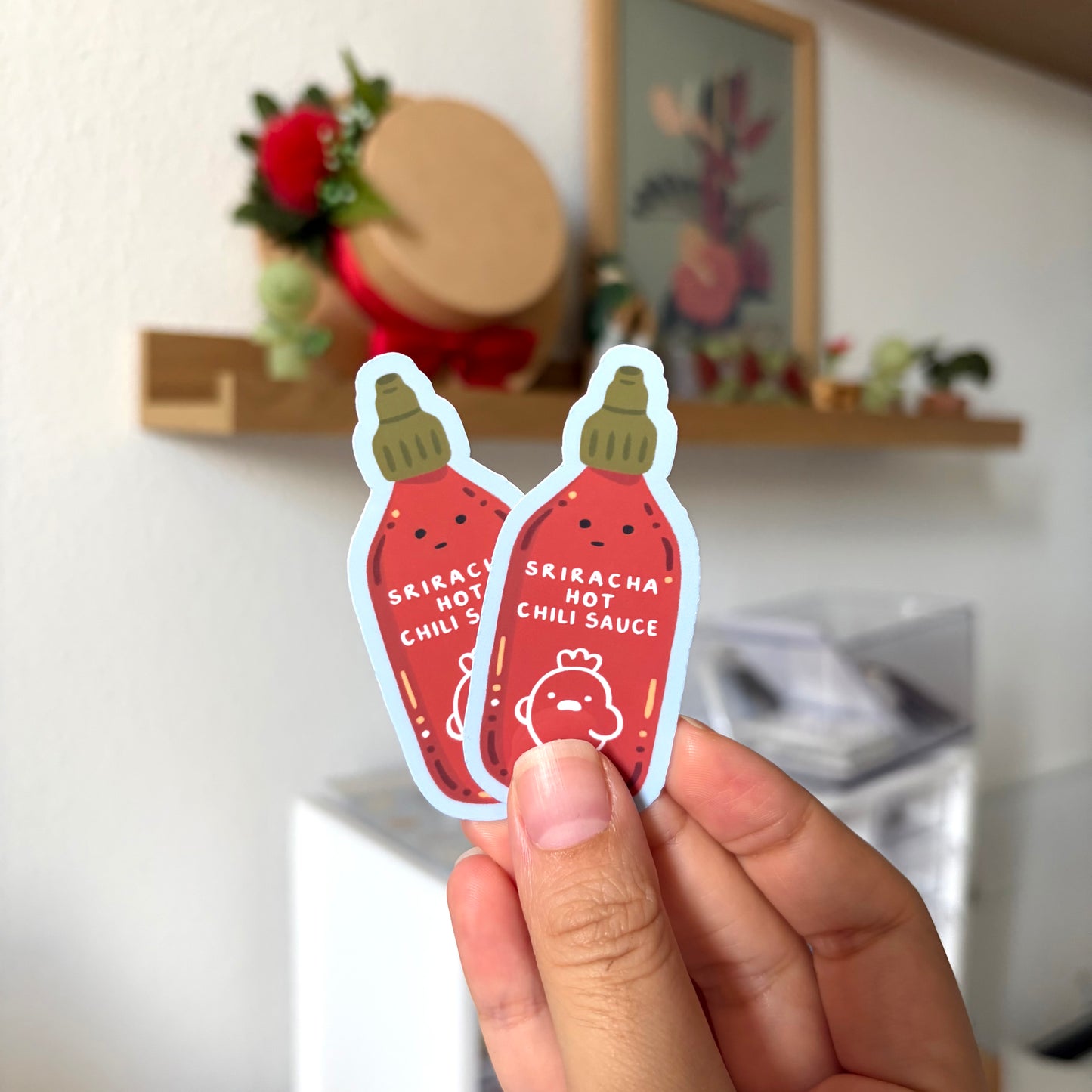 Two Sriracha hot chili sauce stickers held in front of a kitchen background.