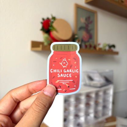 Sticker of a jar labeled 'Chili Garlic Sauce' held by a hand with a blurred indoor background.